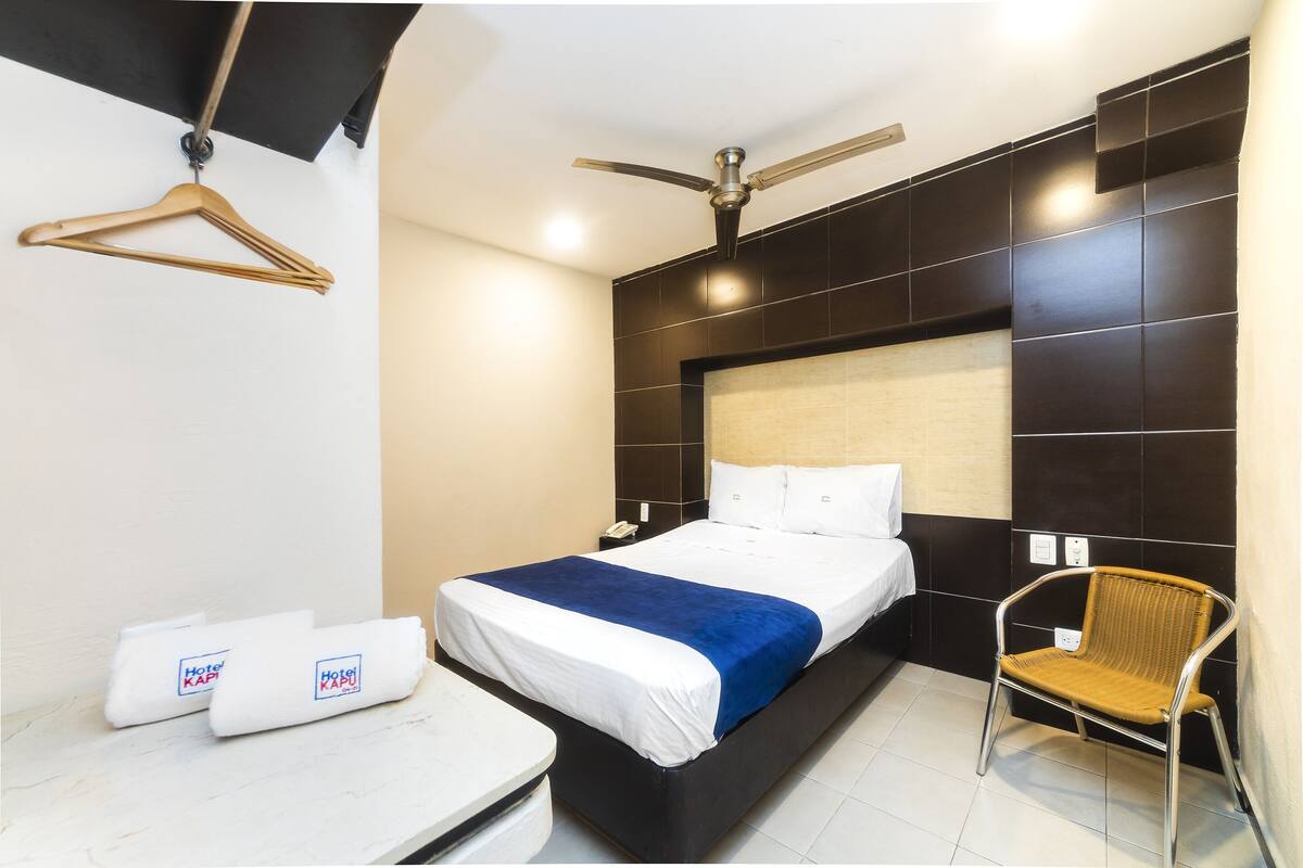 A hotel room with a bed, a chair, a hanging coat rack, and a ceiling fan.