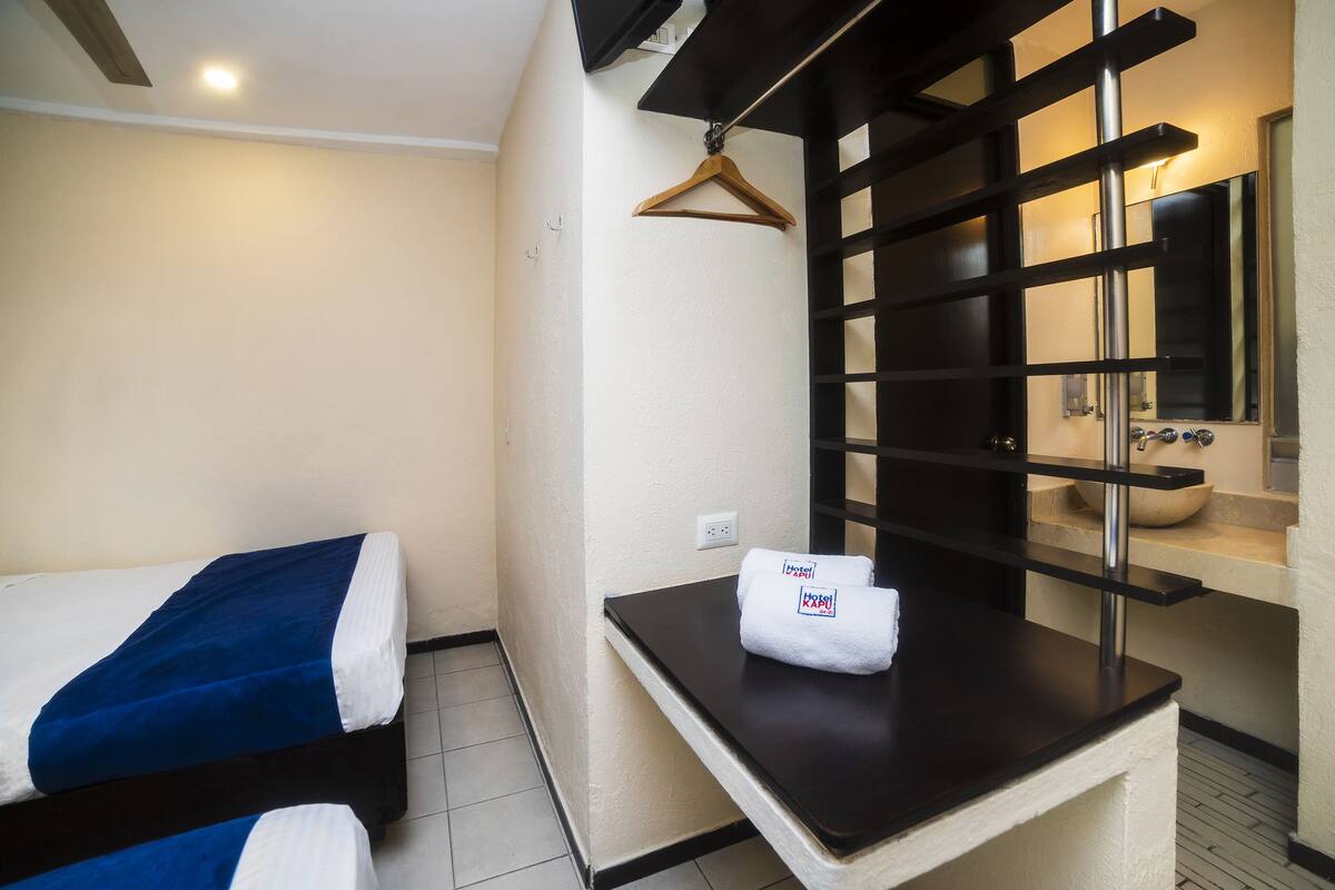 A hotel room with a bed, a small table with a towel, a mirror, and a black shelf unit.
