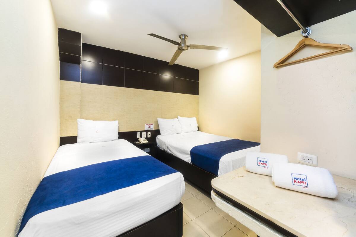 A hotel room with two beds, a ceiling fan, a bedside table, and a hanging coat rack.