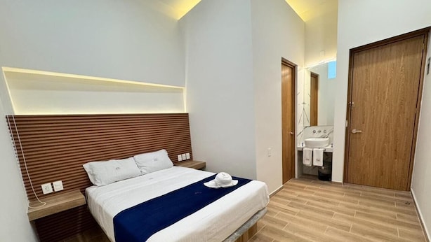 A modern bedroom with a bed, bedside tables, a wooden headboard, a bathroom with a sink and mirror, and a wooden door. - Hotel Kapu Puebla (Puebla)