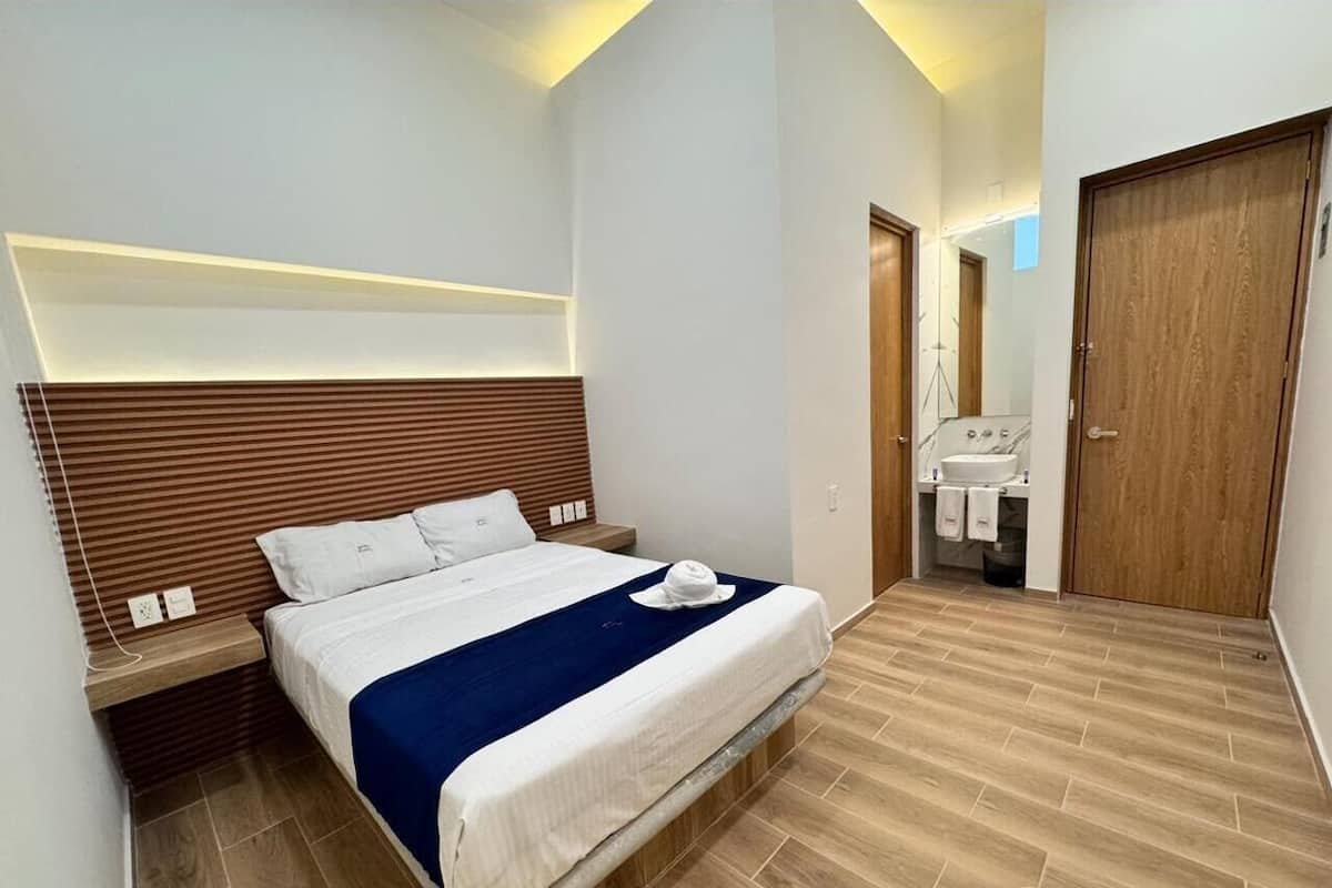 A modern bedroom with a bed, bedside tables, a wooden headboard, a bathroom with a sink and mirror, and a wooden door.