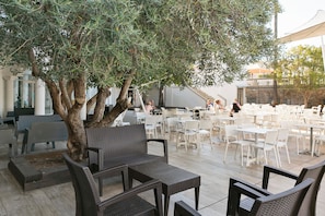 Un espacio exterior con mobiliario de mimbre y un gran árbol. Mesas y sillas dispuestas para comer.