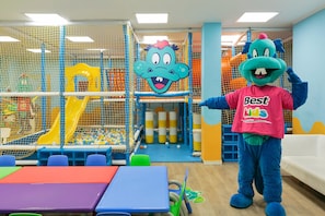 Zona de juegos con tobogán, estructuras de escalada con red y un personaje mascota.