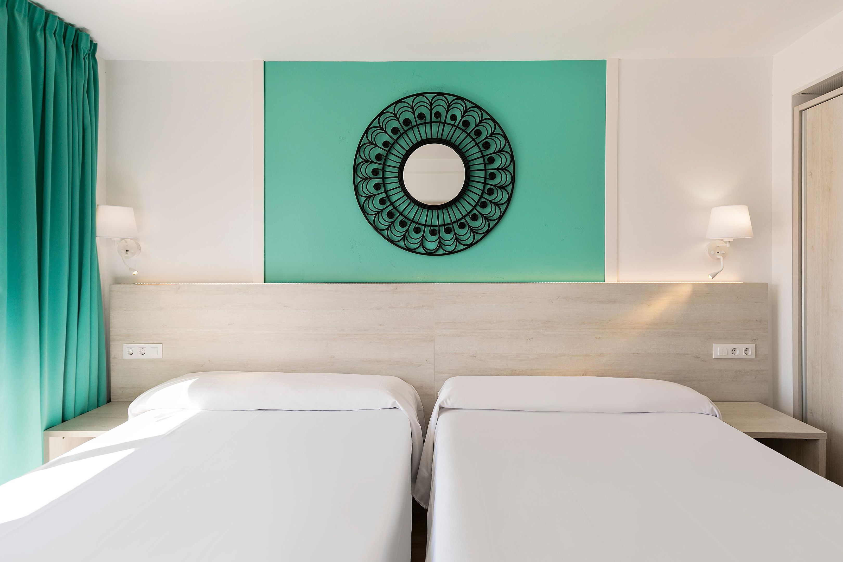 Habitación de hotel con dos camas individuales, una pared de acento color turquesa, un espejo decorativo circular y lámparas de noche.