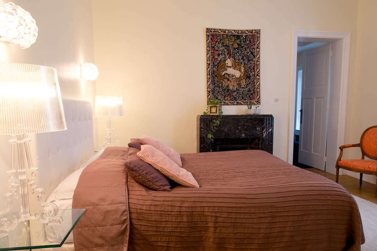 A bedroom with a bed, a glass table, a lamp, a chair, and a fireplace.