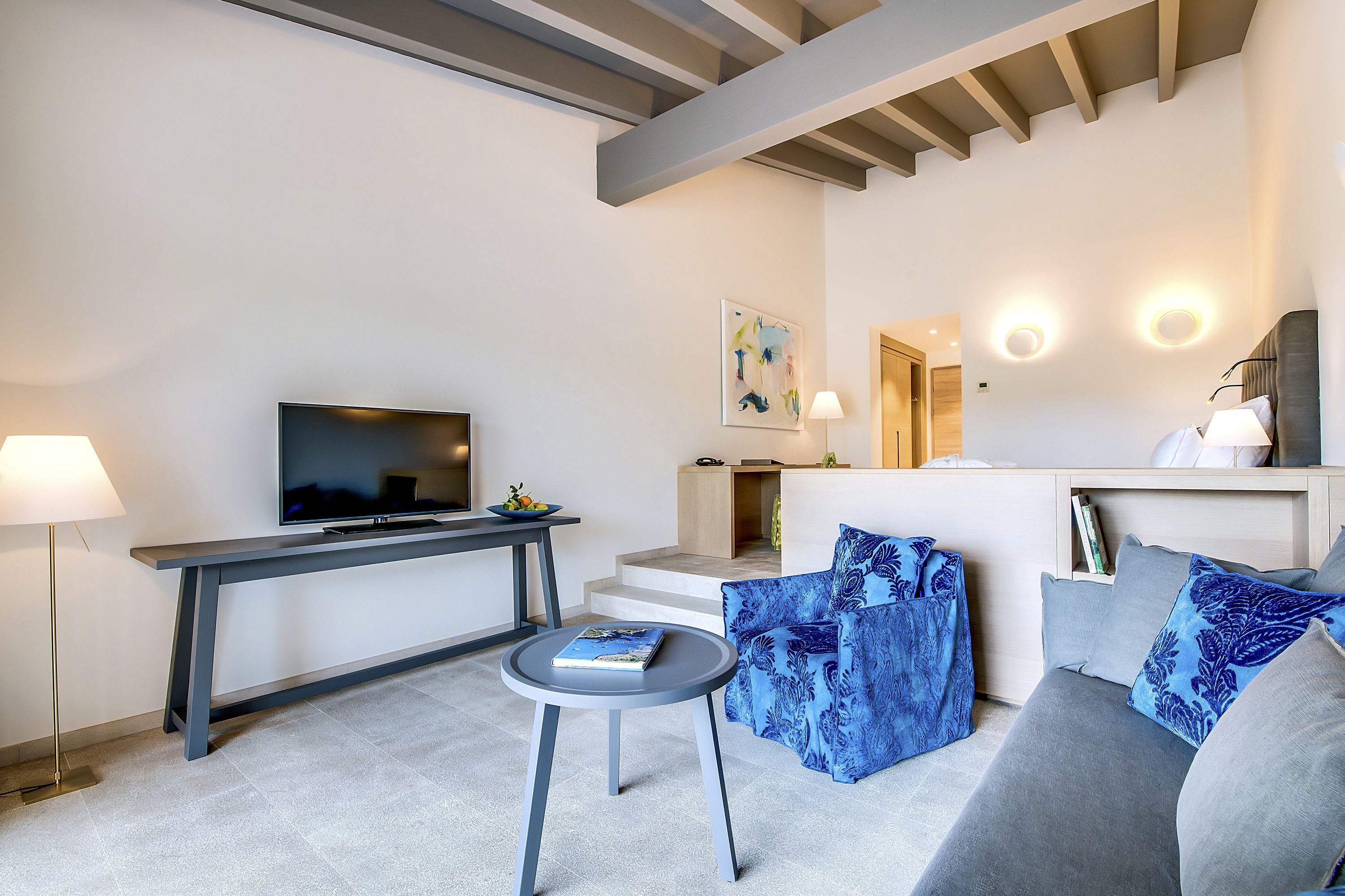 A modern living room with a flat-screen TV, a round table, a sofa with patterned cushions, and a staircase leading to an upper level.