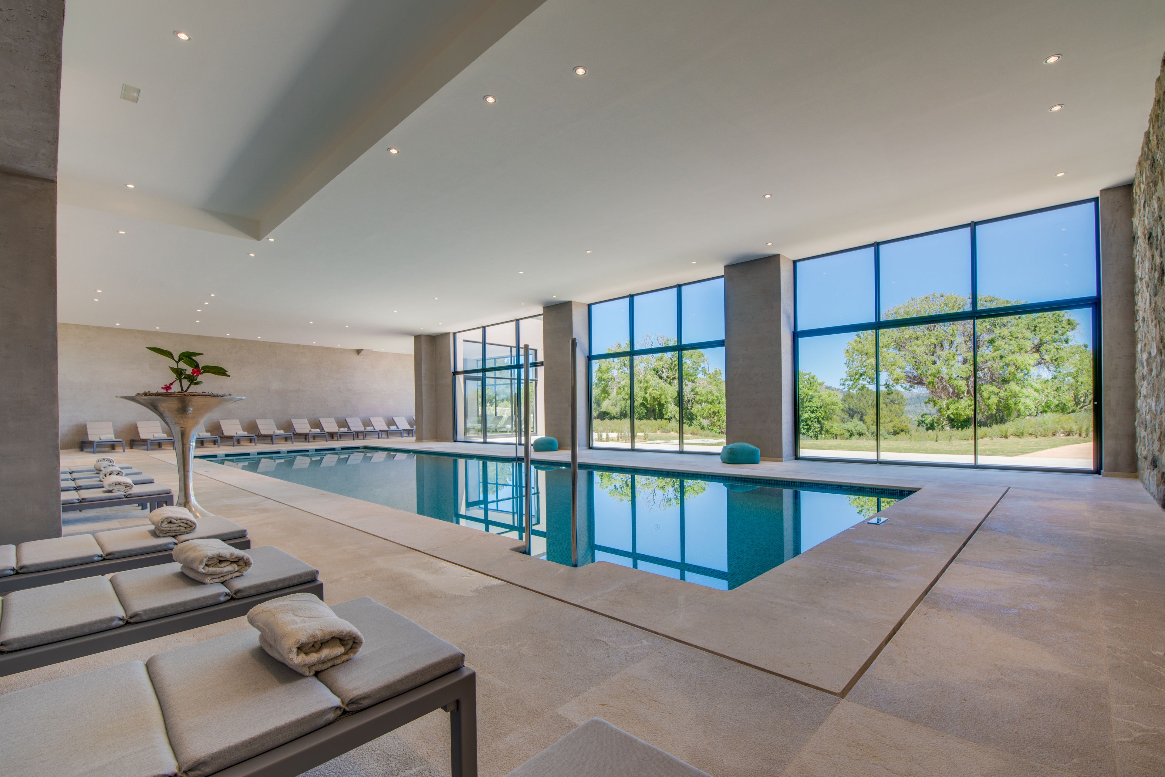 A modern indoor swimming pool with clear glass walls, sun loungers, and a stone-tiled floor.