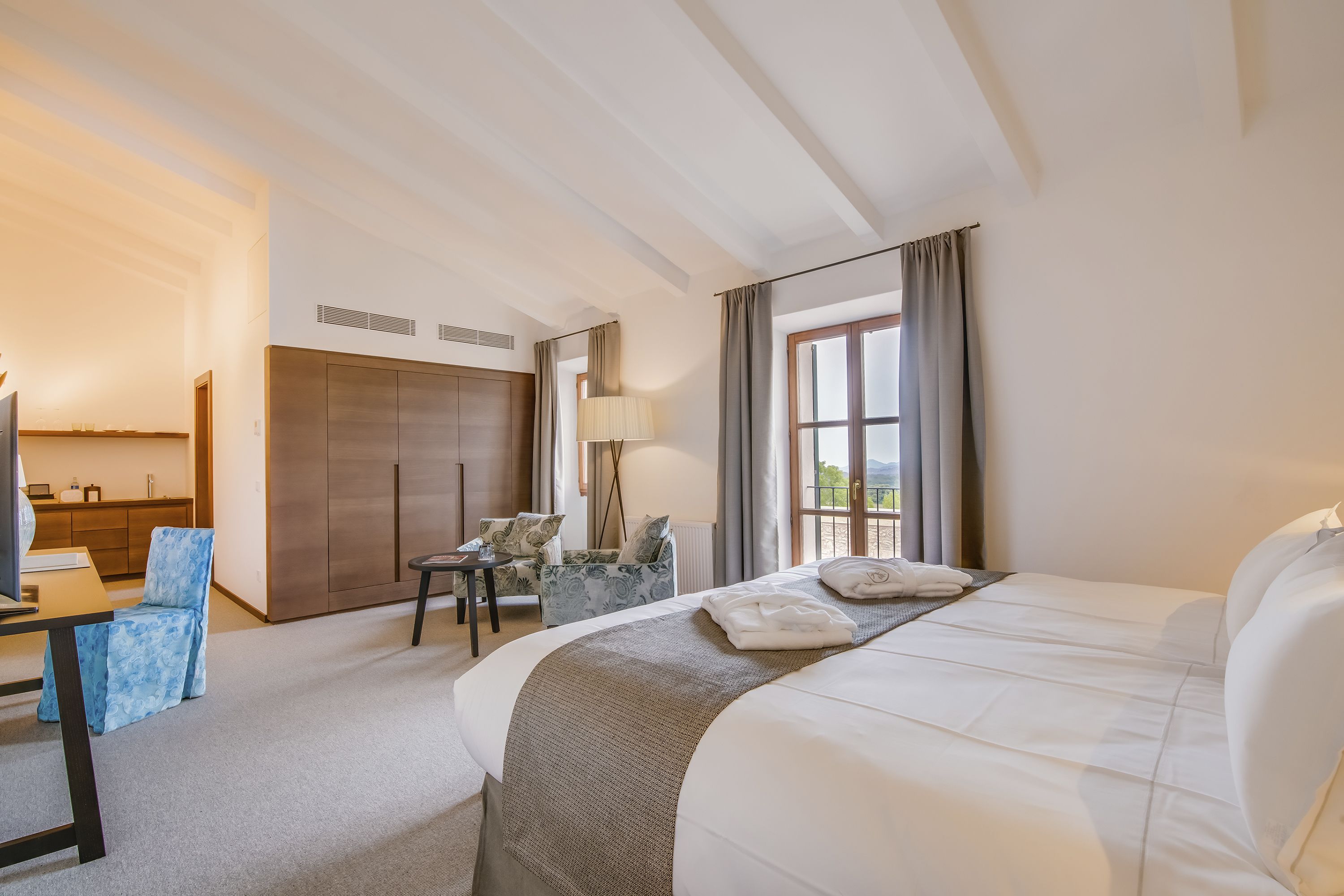A hotel room with a large bed, a desk, a chair, a lamp, and a view of the outdoors.