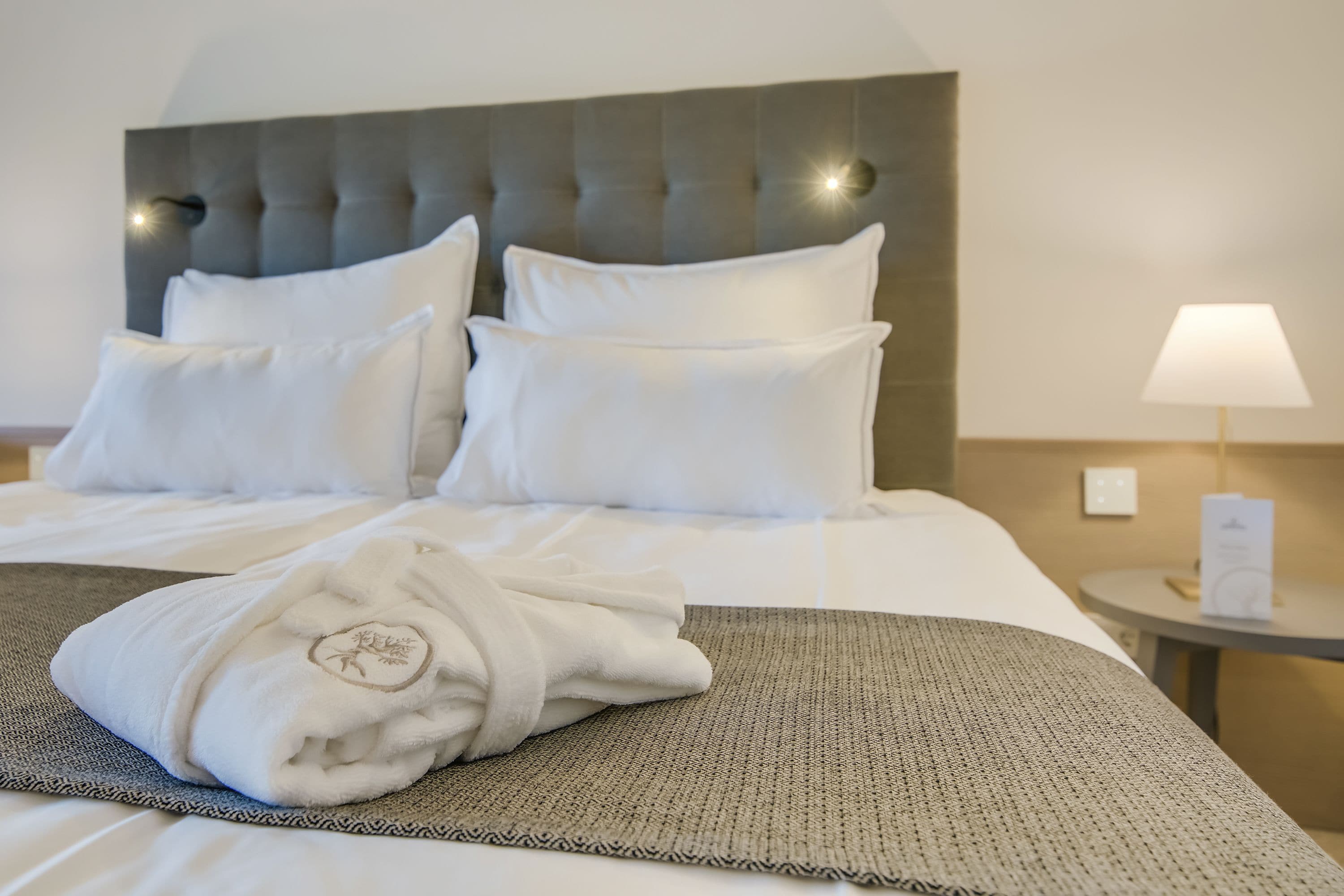 A neatly made bed with a grey headboard, white pillows, and a folded towel on a grey bed runner.
