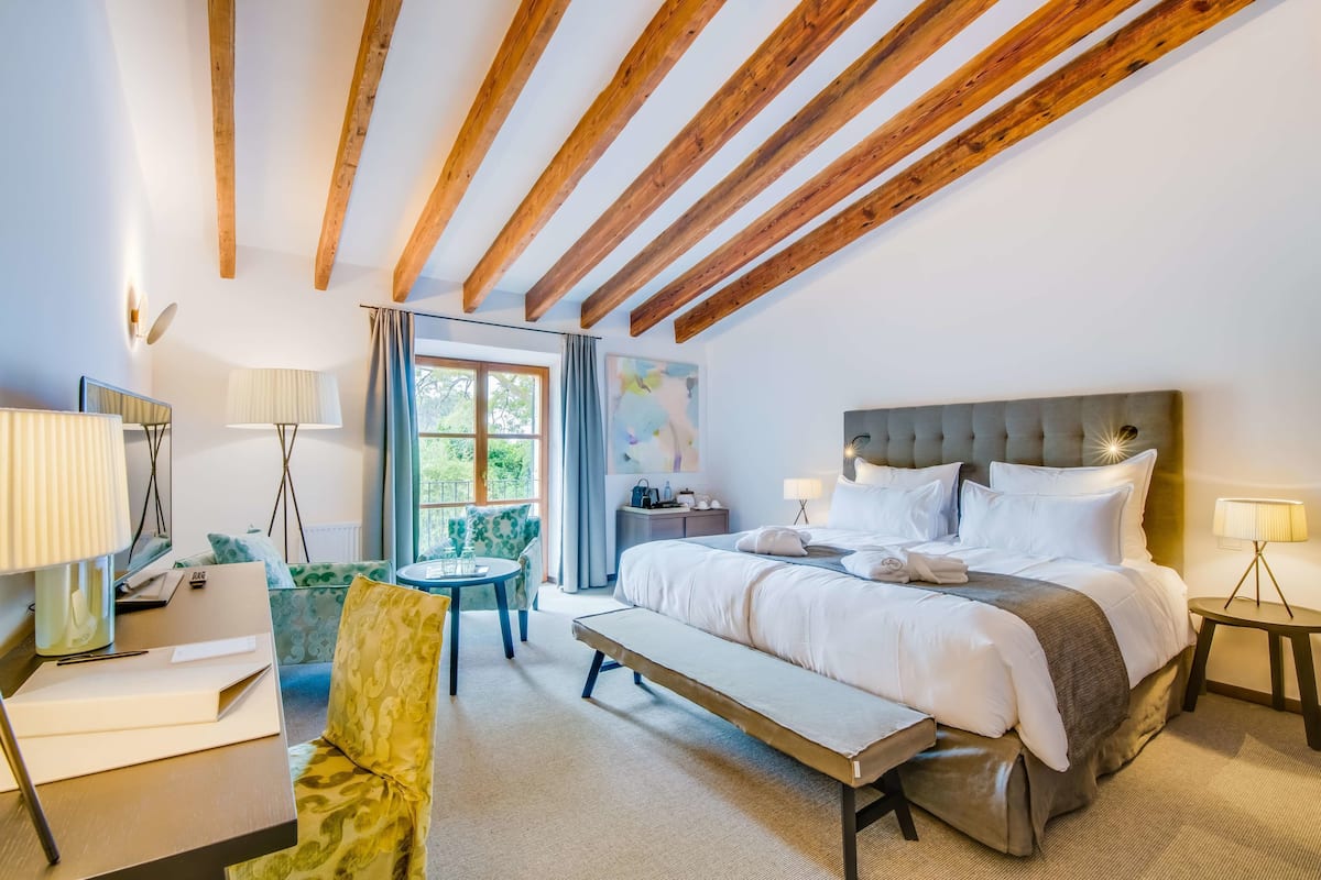 A hotel room with a large bed, a desk, a chair, and a bench. There is a window with curtains, a lamp, and a painting on the wall.