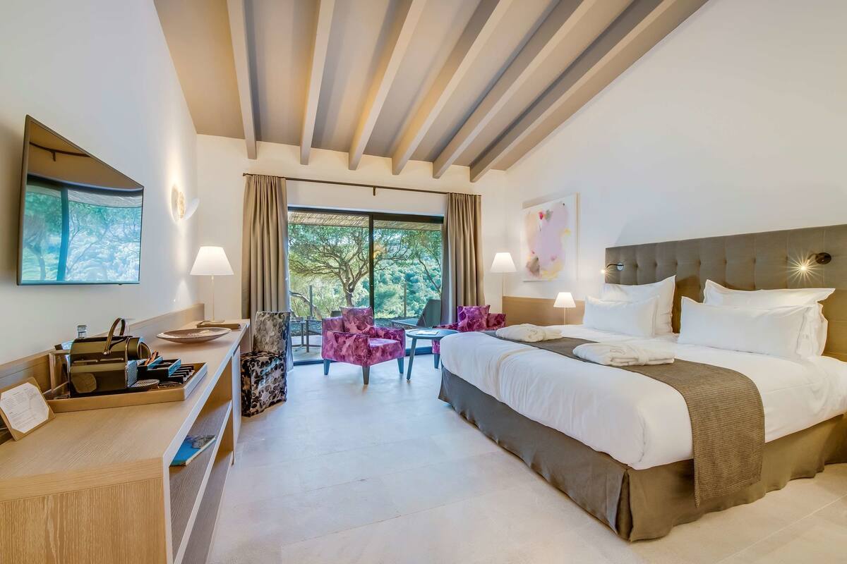 A hotel room with a large bed, a desk with a chair, a small table, and a balcony with chairs.