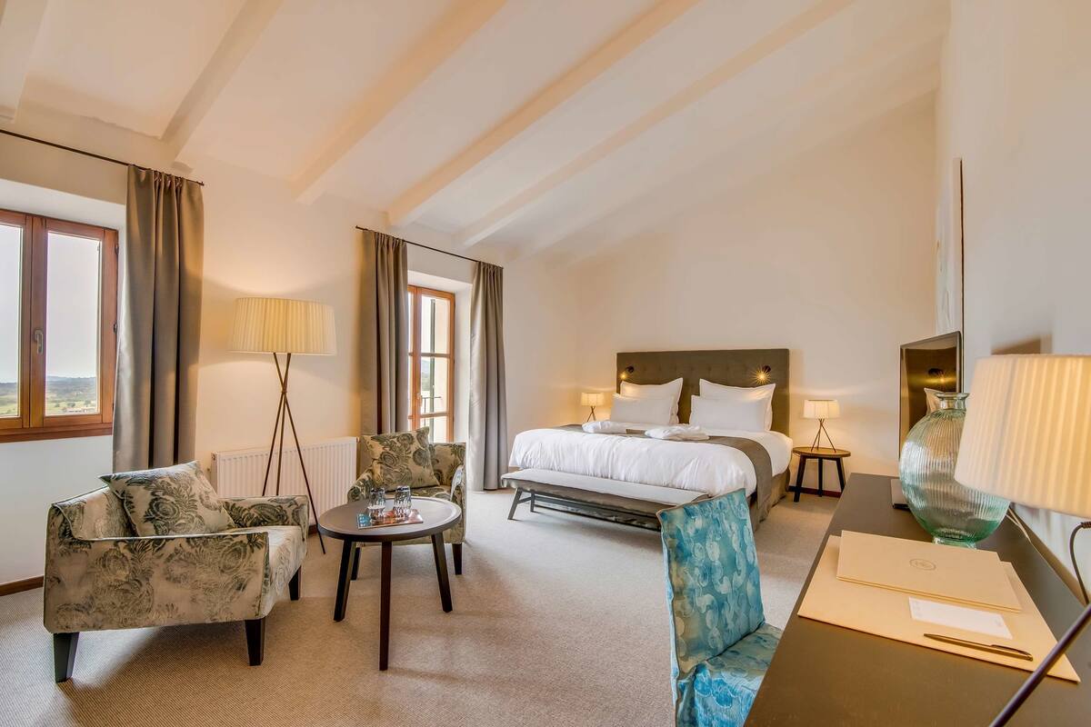 A hotel room with a bed, two armchairs, a small table, a desk, and a lamp.