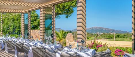 Un espacio de comedor al aire libre con mesas preparadas para dos, con vistas a un paisaje pintoresco con montañas y un viñedo.
