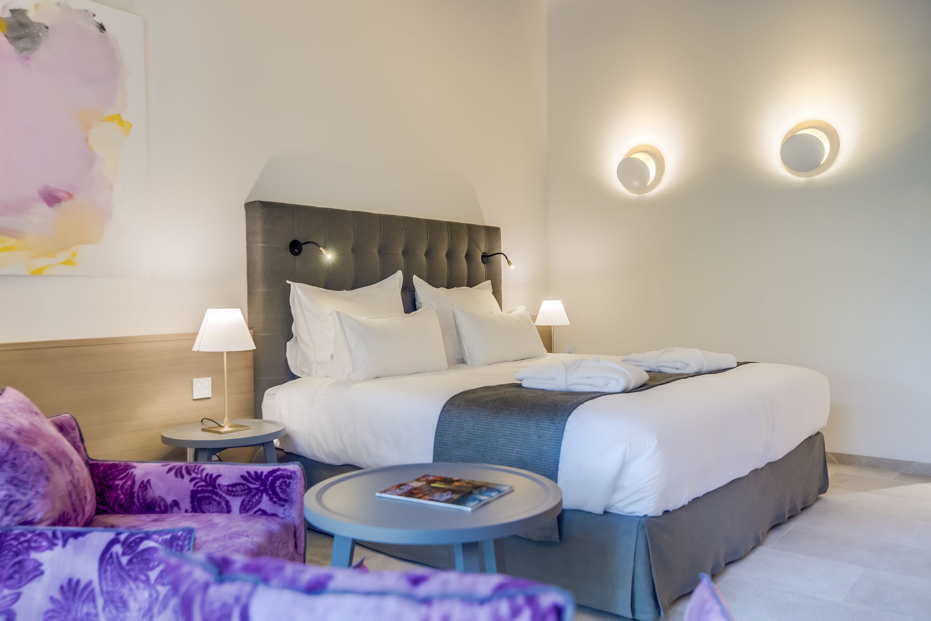 Quarto de hotel moderno com uma cama grande, duas luminárias de cabeceira, uma mesa redonda com uma revista e um sofá floral lilás.