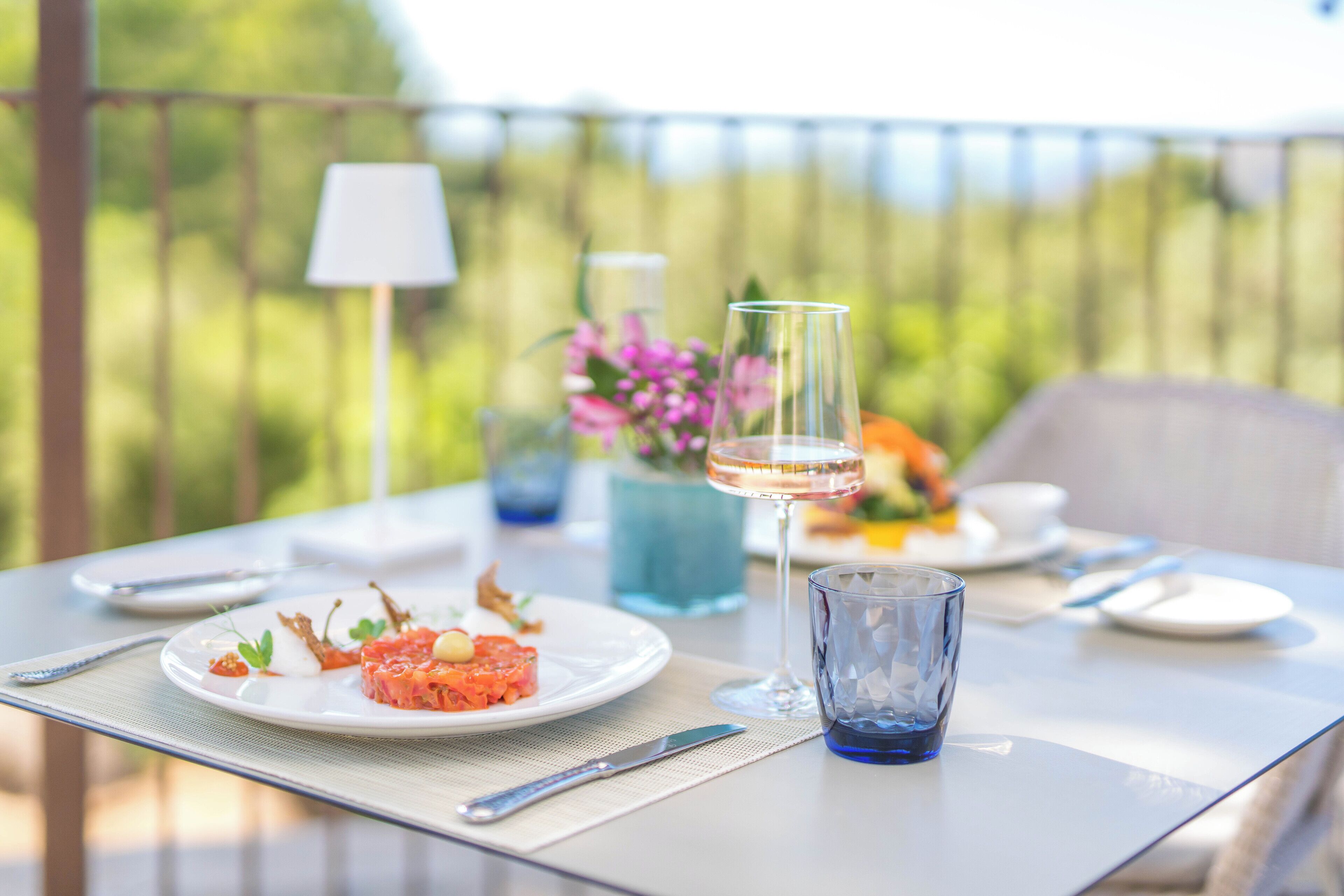 Una mesa dispuesta con un plato de comida, una copa de vino y un jarrón de flores en una terraza al aire libre.