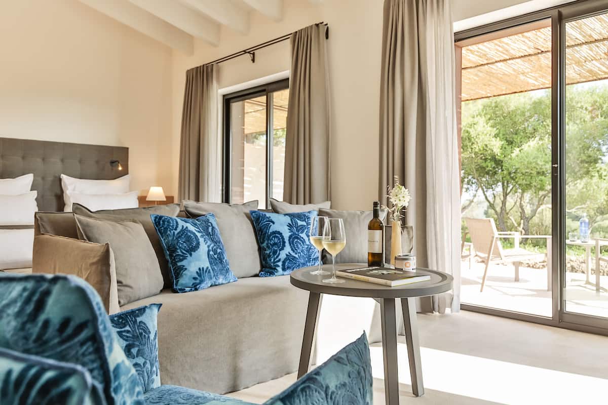 A modern hotel room with a large bed, a sofa, a small round table with a wine glass and a bottle, and a view of the outdoors through large windows.