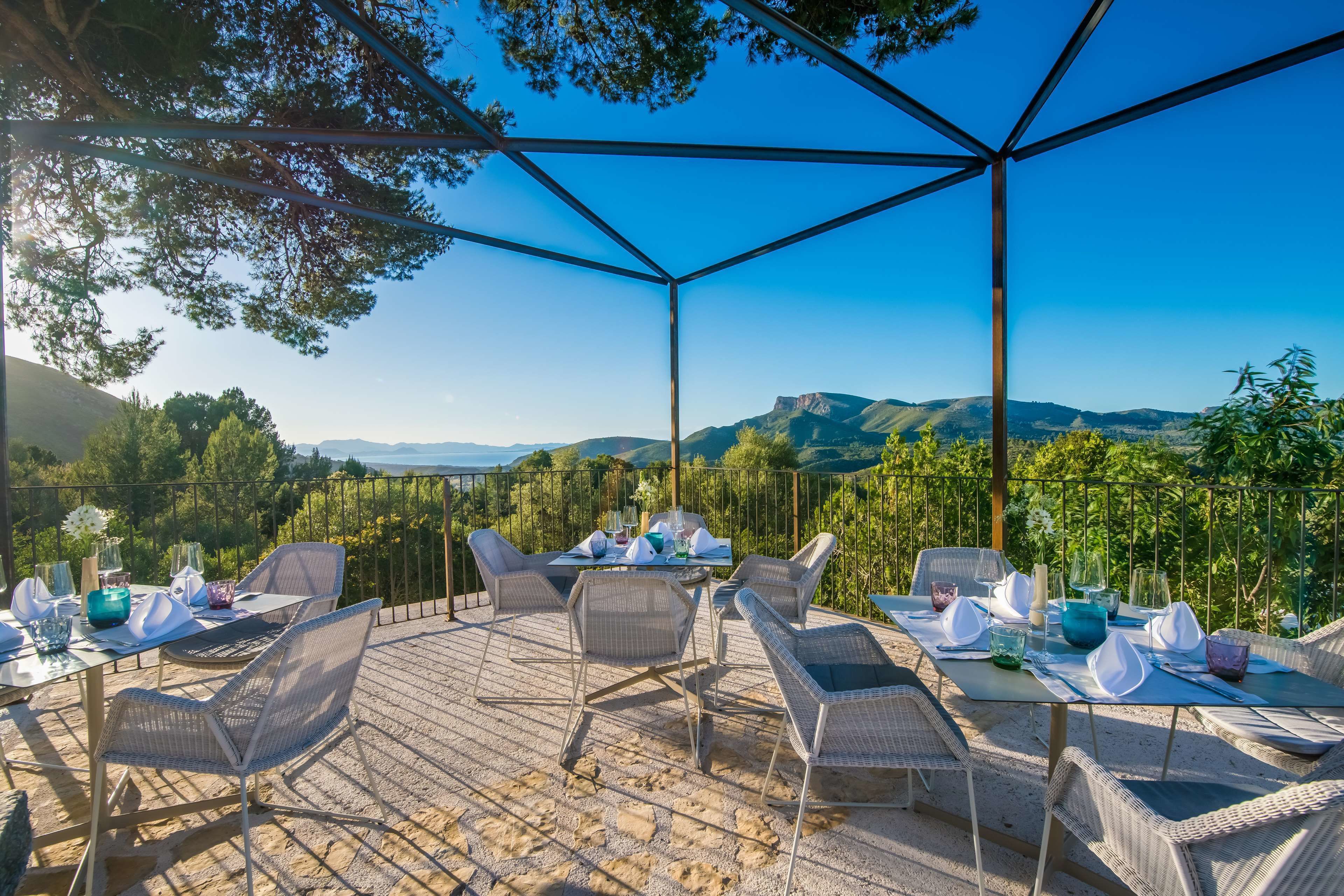 An outdoor dining area with wicker chairs and tables, set against a backdrop of mountains and trees.
