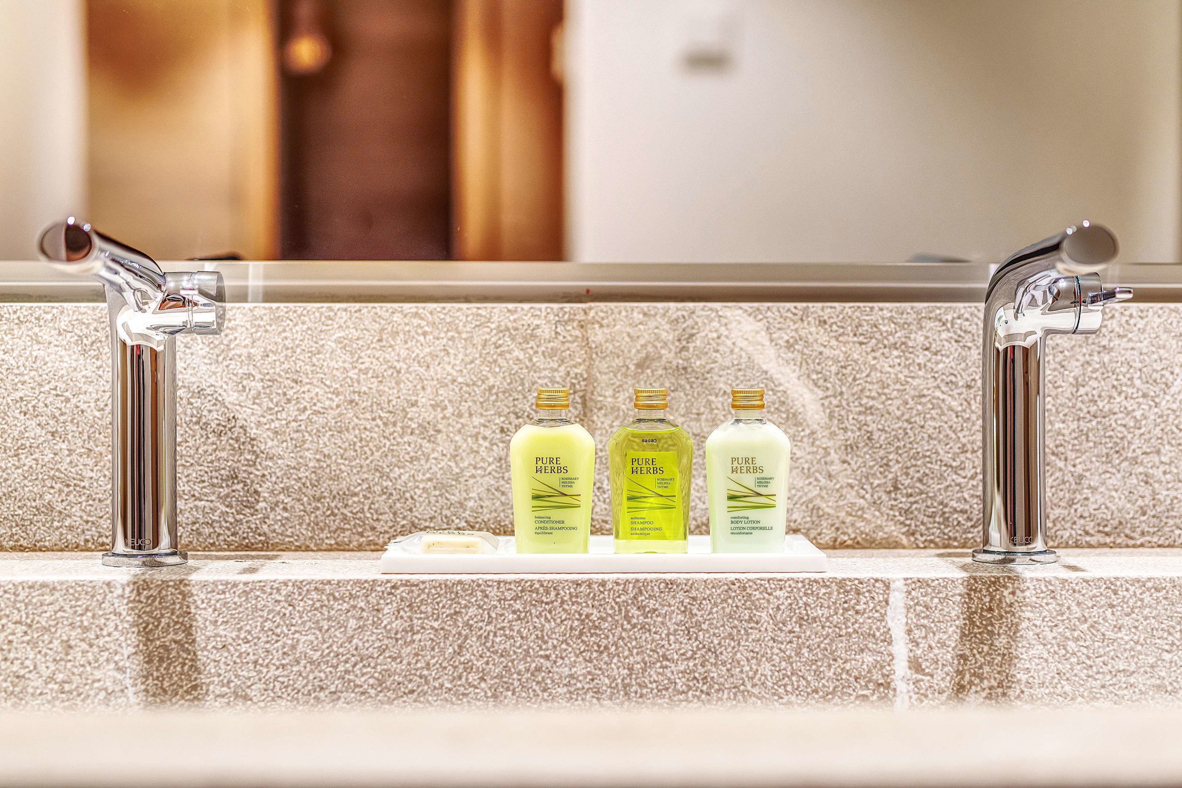 Une salle de bain avec un mur recouvert de carreaux beiges, un robinet chromé et trois flacons de lotion pour le corps de la marque Pure Herbs, posés sur un porte-savon blanc.