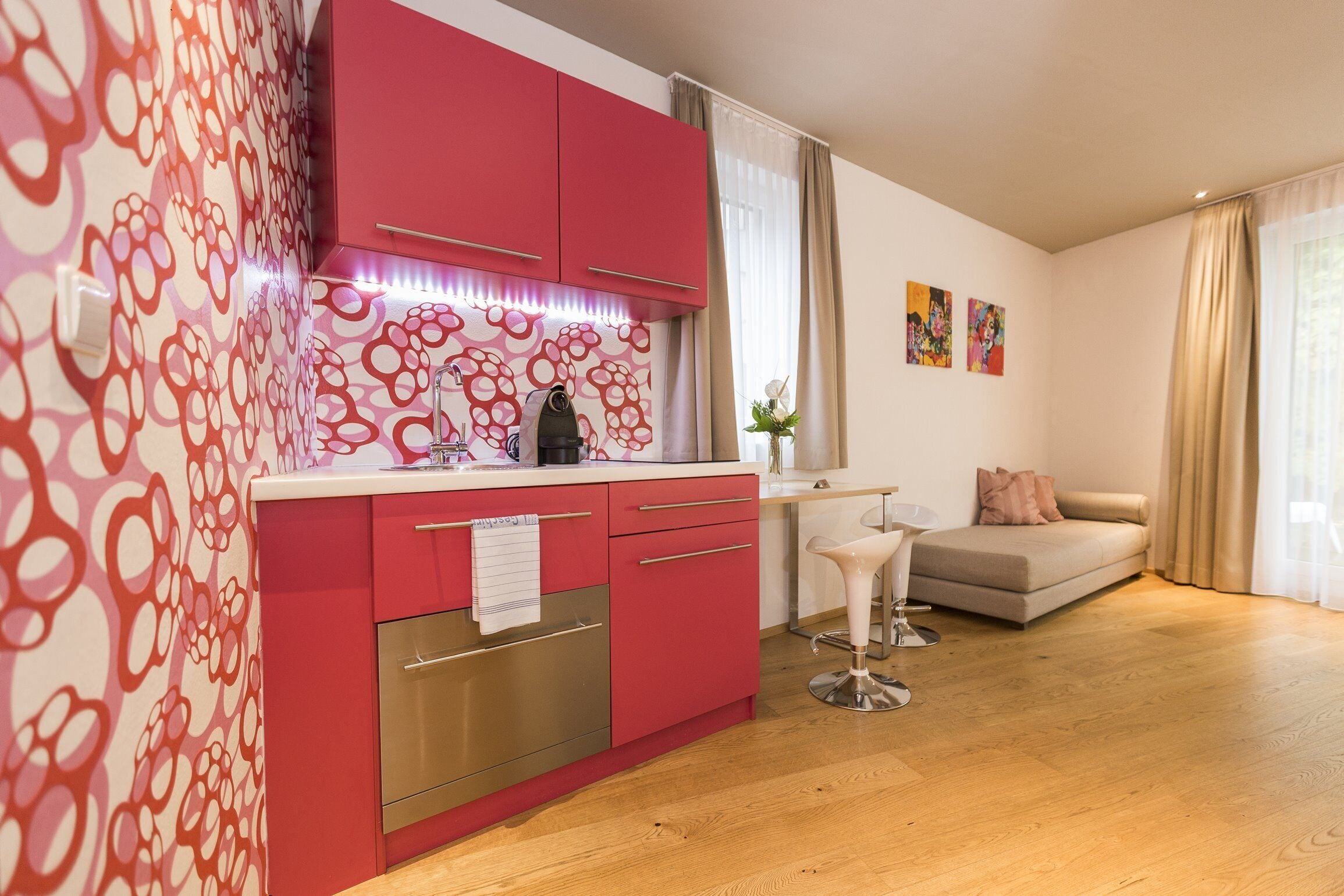 Eine moderne Küche mit roten Schränken, einem Edelstahlbackofen und einer weißen Arbeitsplatte. Im Wohnbereich befindet sich ein beiges Sofa und ein weißer Barhocker.