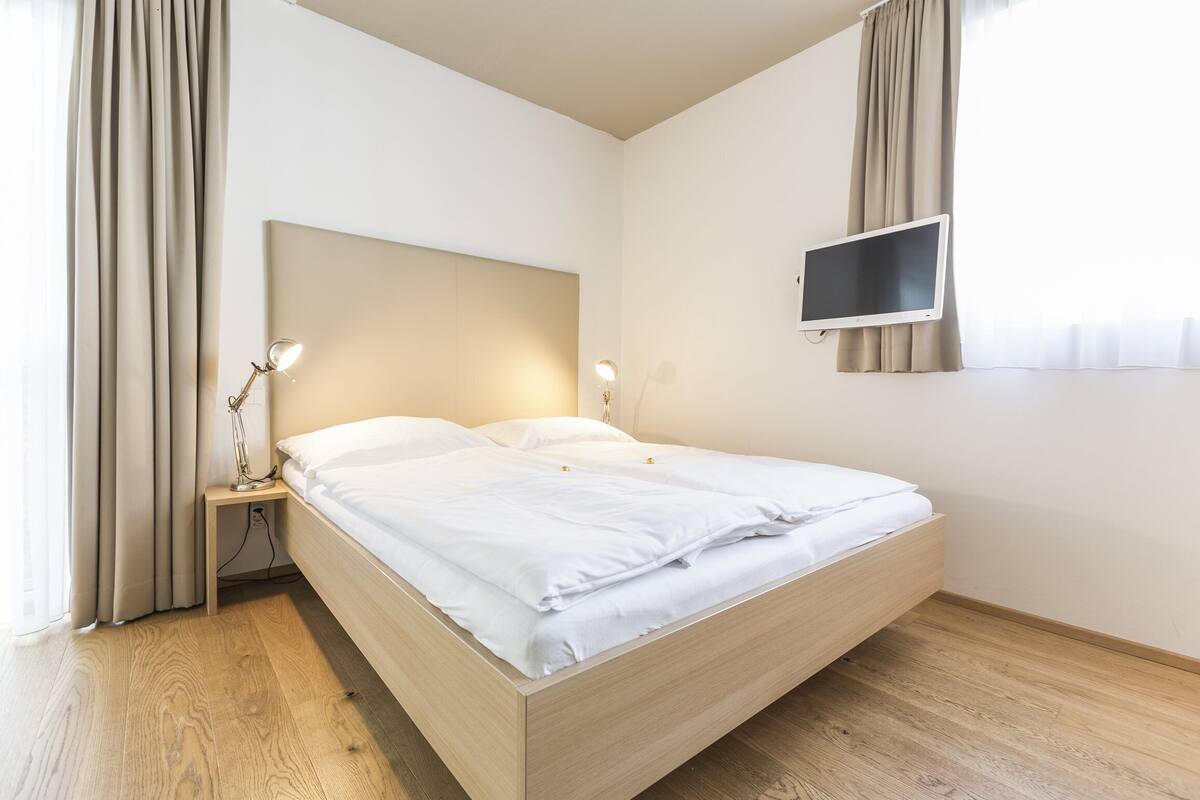 Um quarto moderno com uma cama de madeira, uma mesa de cabeceira com uma luminária, uma televisão de tela plana na parede e uma janela com cortinas.