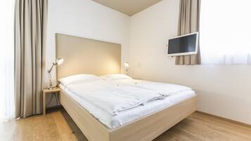 A modern bedroom with a wooden bed, a bedside table with a lamp, a flat-screen TV mounted on the wall, and a window with curtains.