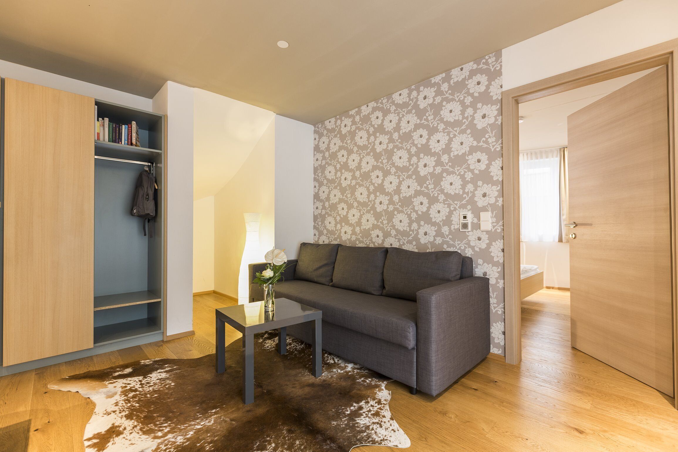 Ein modernes Wohnzimmer mit einem grauen Sofa, einem Glastisch und einem Kuhfellteppich. Ein Bücherregal mit Büchern und einer Jacke, dazu floraler Wandbelag.