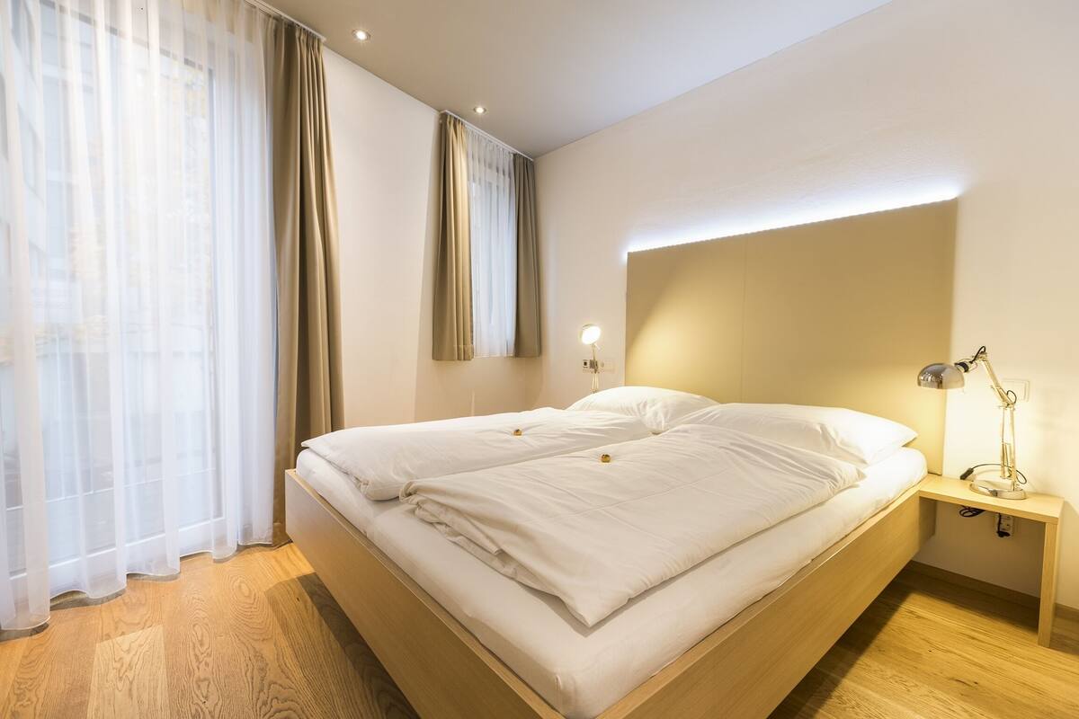 Quarto moderno com cama de madeira, roupa de cama branca, mesa de cabeceira com abajur e cortinas leves.