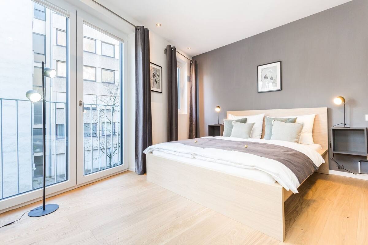 Quarto moderno com uma cama grande, mesa de cabeceira, televisão e vista da cidade através de uma janela ampla.