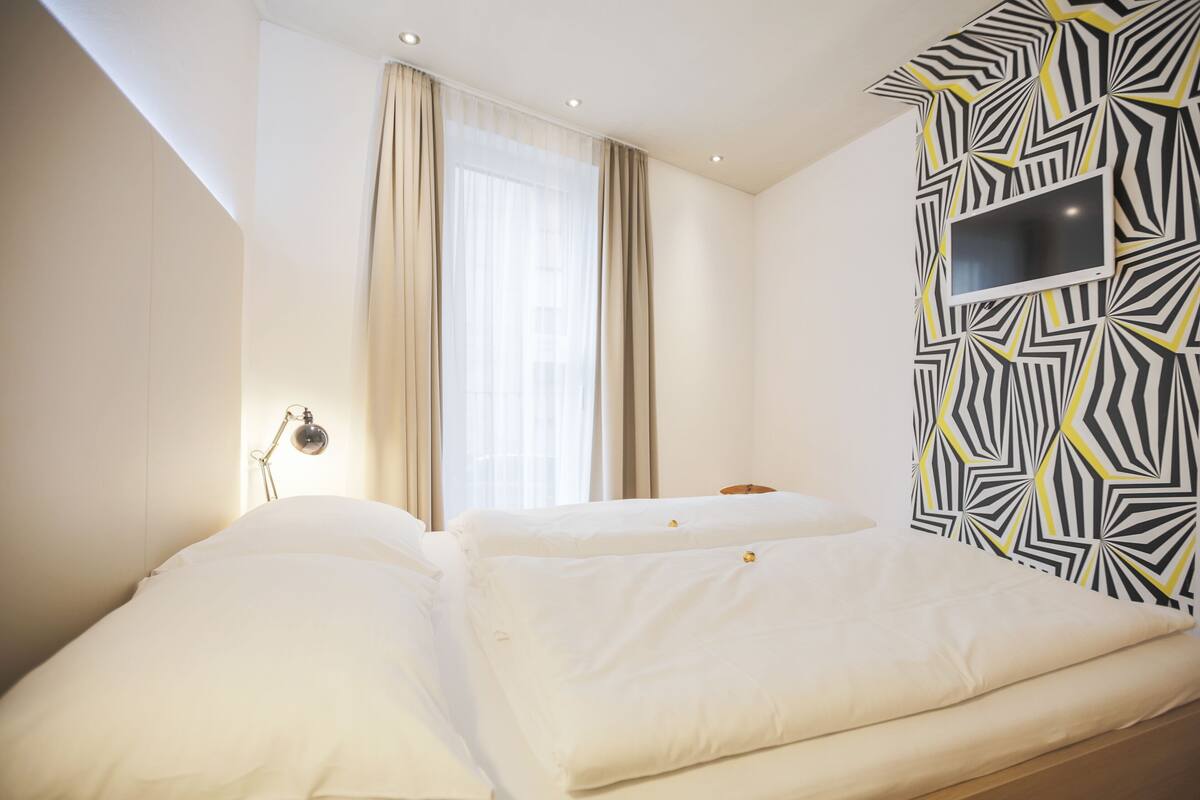 Quarto de hotel com duas camas, uma janela grande com cortinas, uma TV fixada na parede e um abajur.