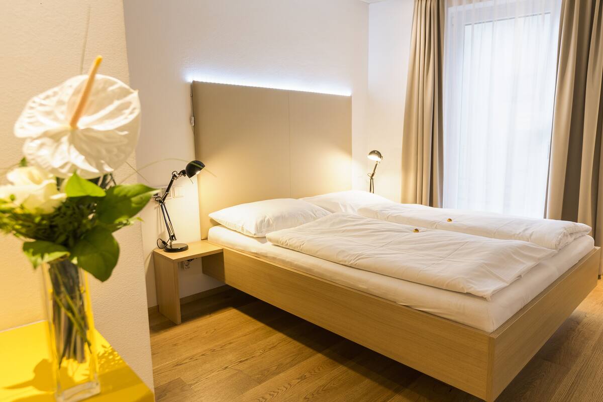 Quarto de hotel moderno com cama de madeira, luminárias de cabeceira, uma mesinha e um vaso com flores.