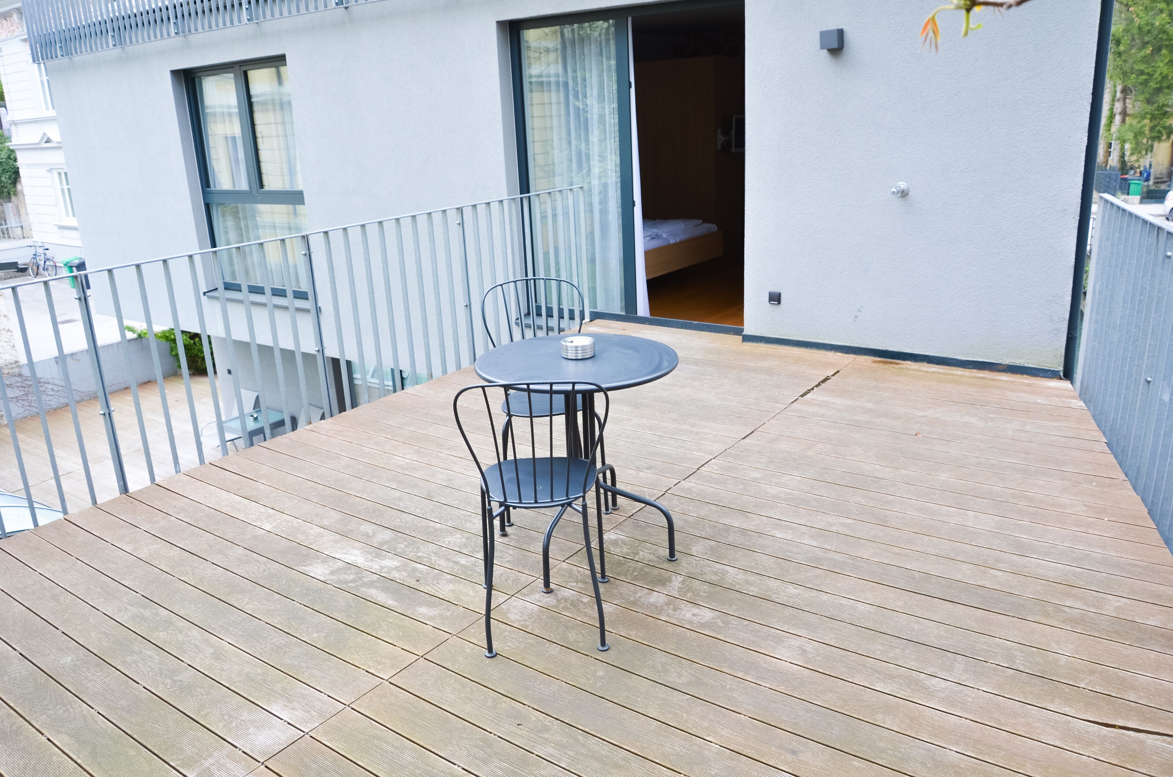 Una terraza de madera con una mesa redonda y dos sillas, una puerta corrediza de vidrio que da a un dormitorio y un edificio blanco con balcones.