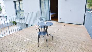 Una terraza de madera con una mesa redonda y dos sillas, una puerta corrediza de vidrio que da a un dormitorio y un edificio blanco con balcones.