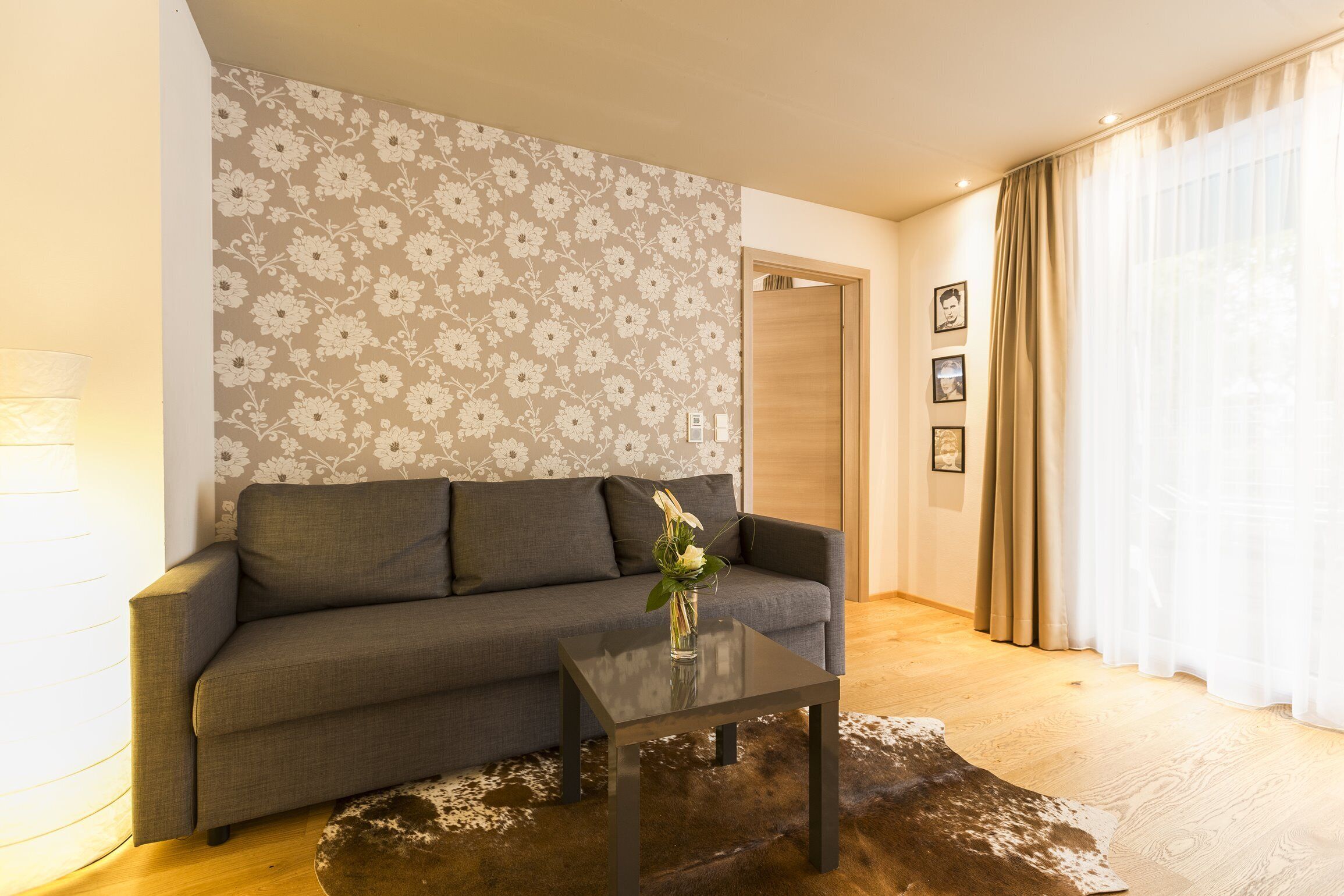 A living room with a sofa, a coffee table, a floral wallpaper, and a wooden floor.