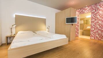 Een moderne slaapkamer met een houten bed, een aan de muur bevestigde televisie en een badkamer die door een open deur zichtbaar is.