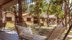 A hammock chair with a patterned cushion in a covered outdoor area with tiled flooring, surrounded by palm trees and a building with a tiled roof. - Orla Náutica Pousada (Barreirinhas)