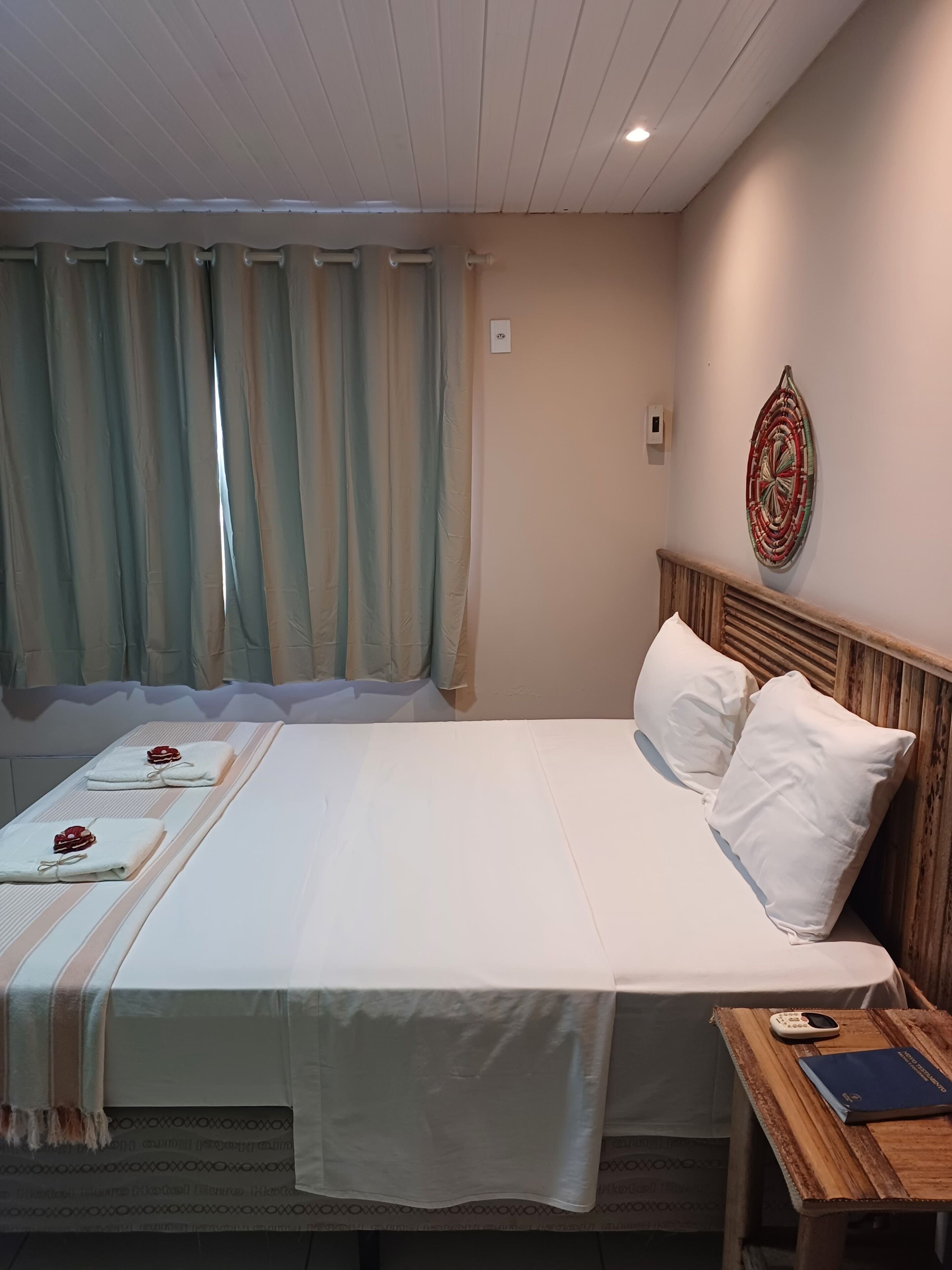 A neatly made bed with white linens and two pillows, a wooden nightstand with a book and a phone, and a wall-mounted decorative piece.