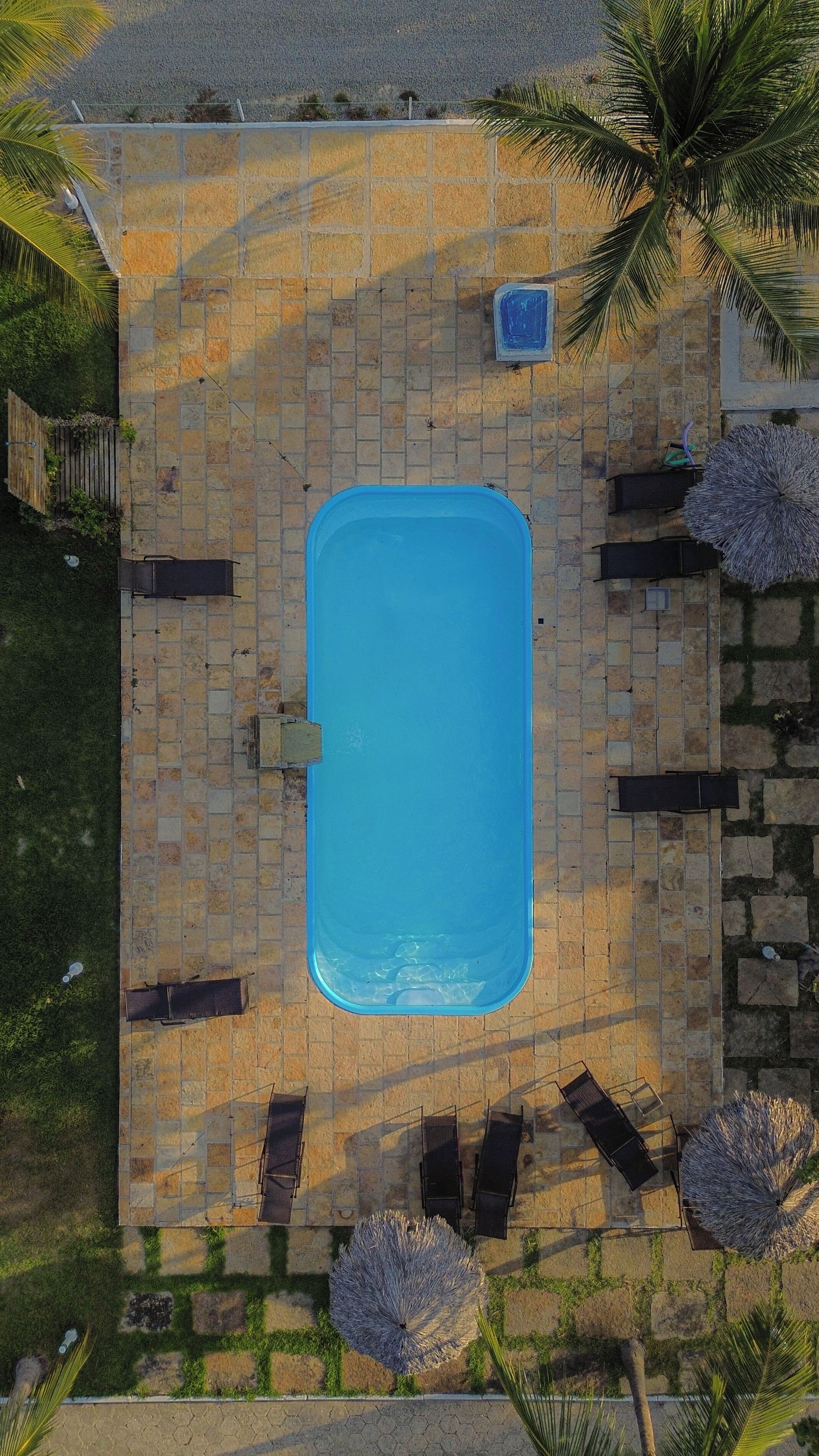An aerial view of a rectangular swimming pool surrounded by stone tiles, with a few umbrellas and chairs around it.