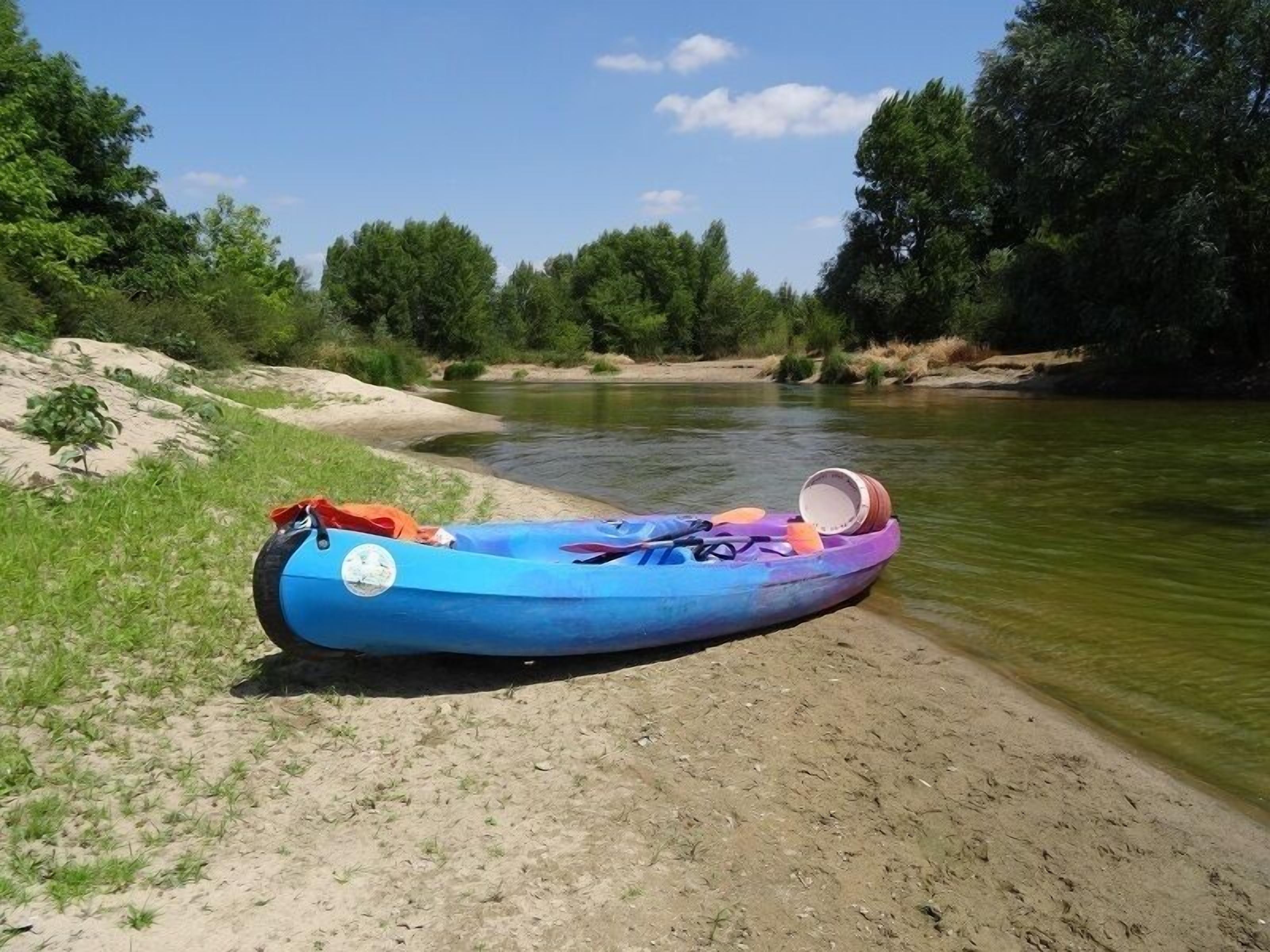 Kayak azul con remo y casco sobre la orilla arenosa de un río.