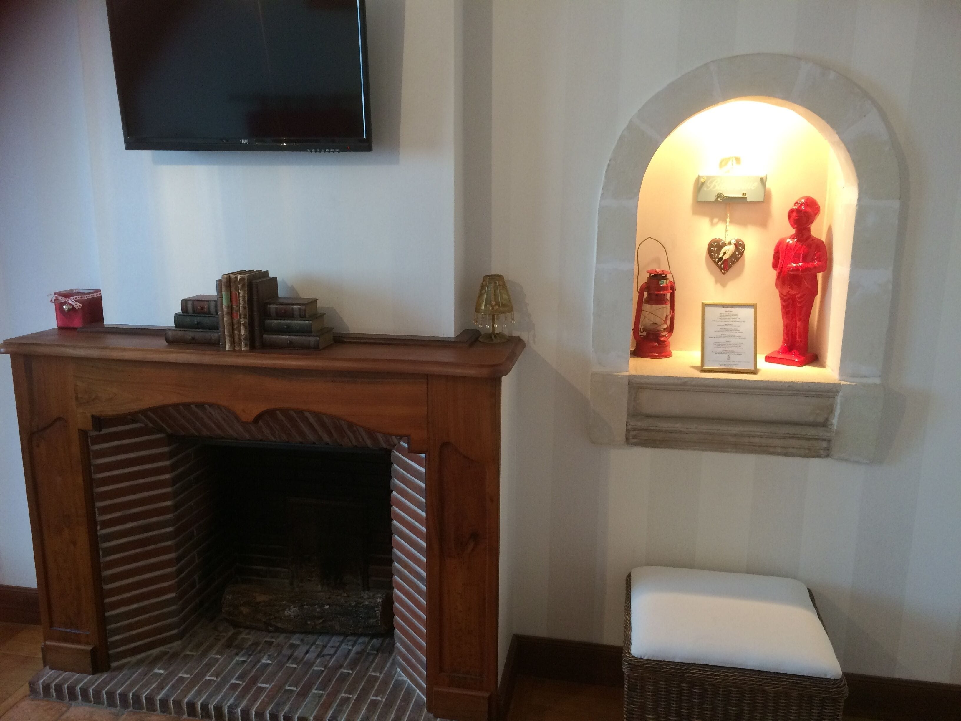 Una chimenea con repisa que muestra libros y un farol rojo, un televisor montado arriba y un pequeño banco con cojín.
