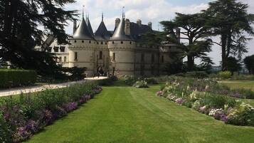 Un edificio storico con una facciata simmetrica, diverse torri e un giardino curato con un prato centrale e aiuole fiorite.