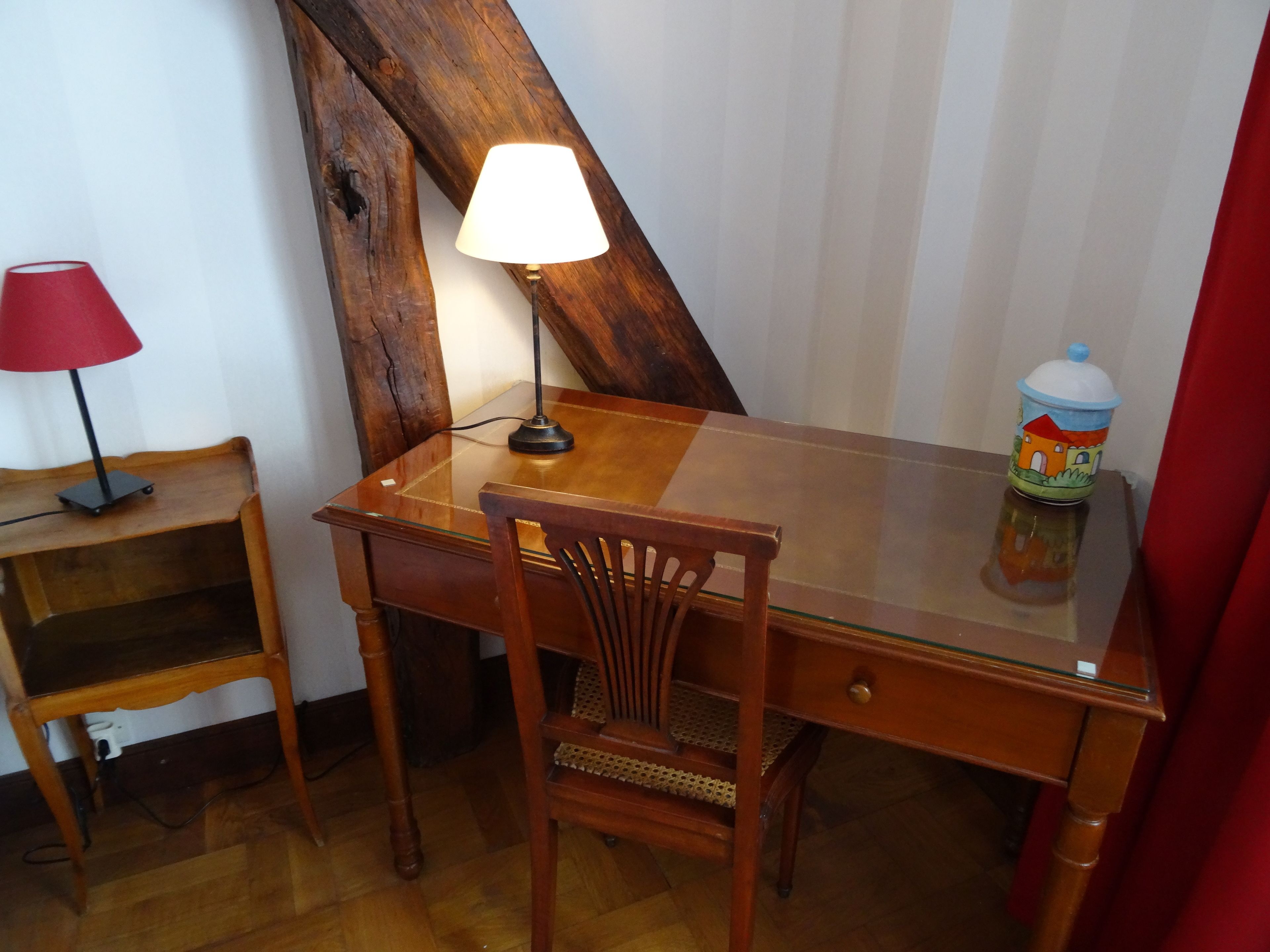 Un bureau en bois surmonté d’une plaque de verre, une lampe rouge, une chaise et un récipient coloré posé sur le bureau.