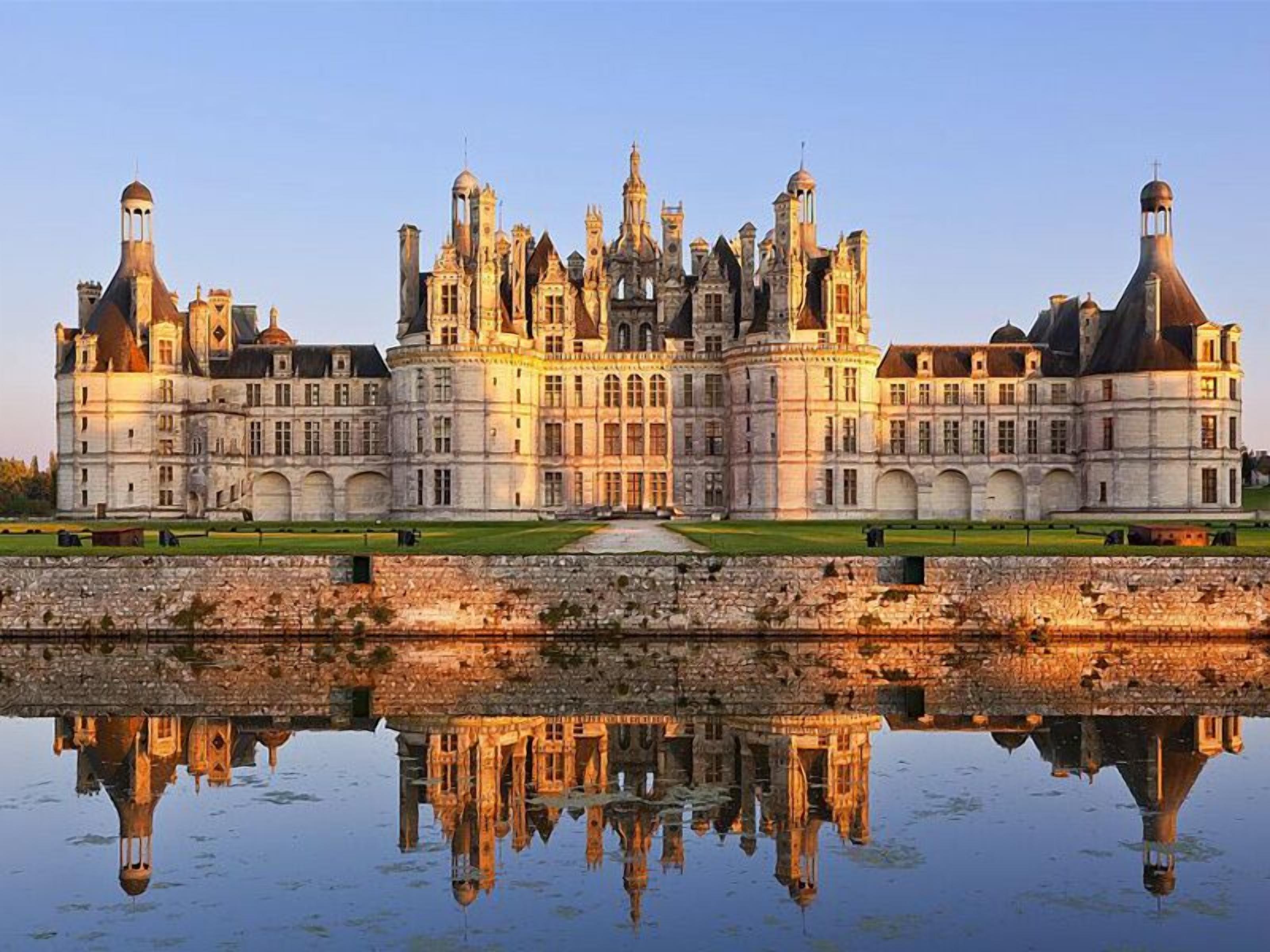 Un edificio grande e histórico con múltiples torres y una fachada simétrica, reflejado en una superficie acuática tranquila.