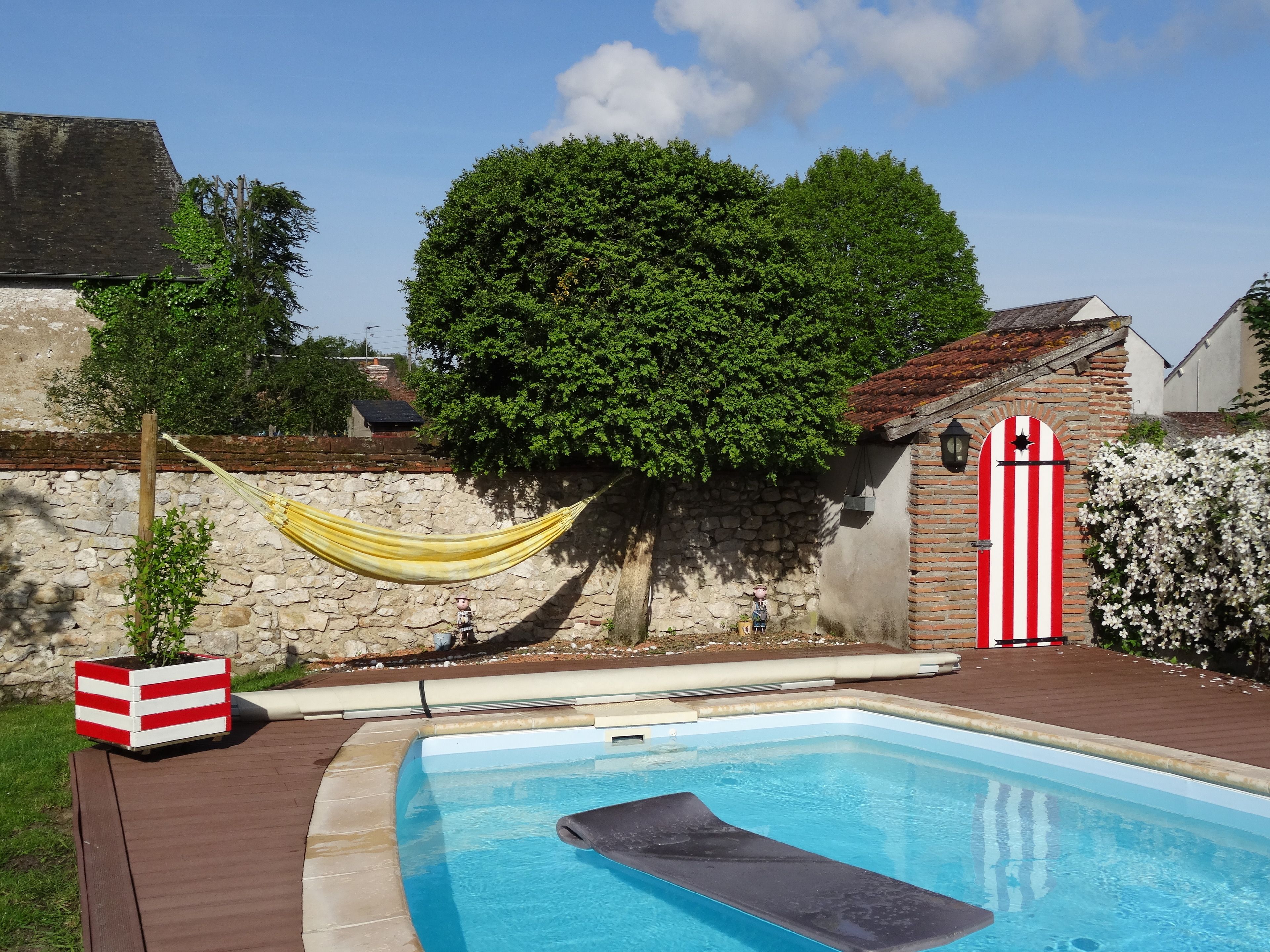 Una piscina con muelle de madera, una hamaca, un muro de piedra y un portón a rayas rojas y blancas.