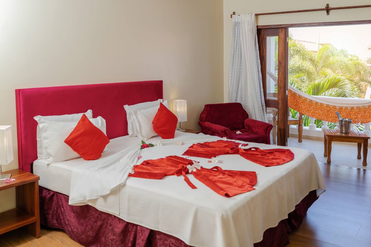 Une chambre d’hôtel avec un lit, une table de chevet, un fauteuil rouge, une table avec une lampe et une vue sur l’extérieur.