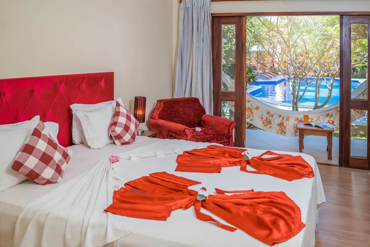 Une chambre d’hôtel avec une tête de lit rouge, du linge de lit blanc et une vue sur une piscine et la verdure environnante.