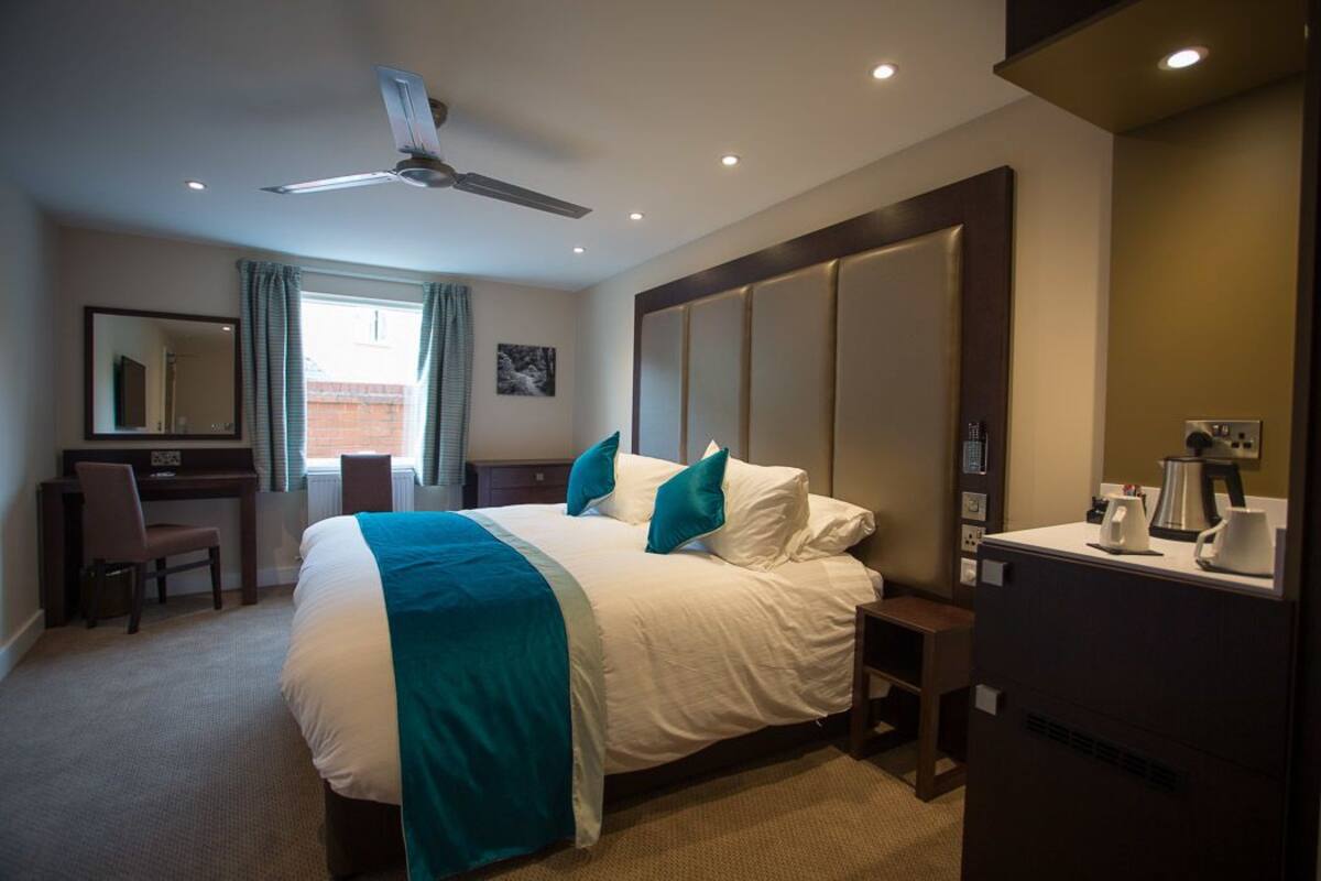 A hotel room with a bed, a desk, a chair, a coffee machine, and a ceiling fan.