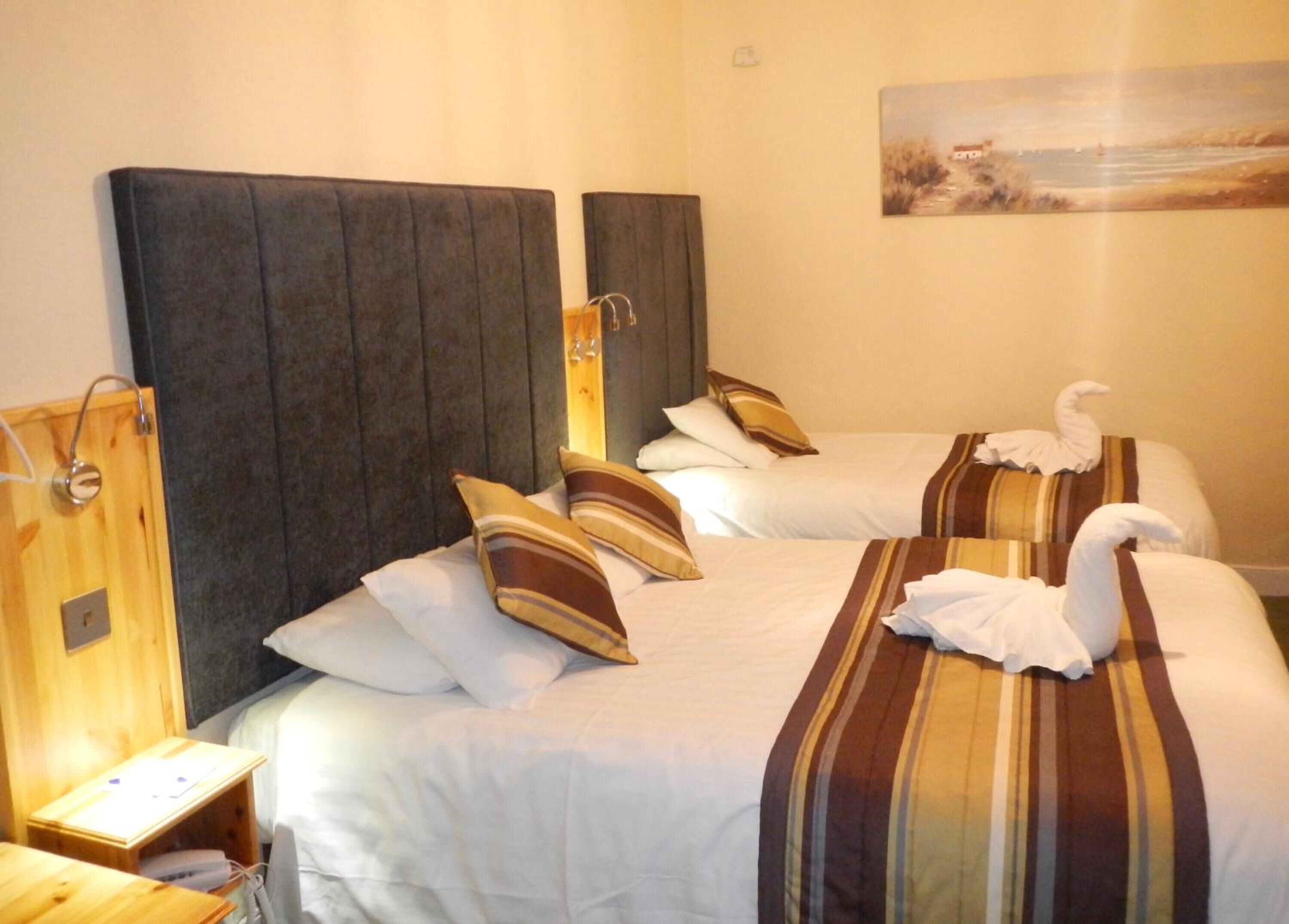 Habitación de hotel con dos camas, cada una con almohadas a rayas y una toalla con forma de cisne. Hay una mesita de noche de madera con una lámpara y un cuadro en la pared.