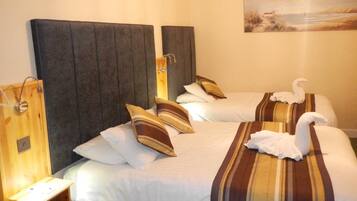 Habitación de hotel con dos camas, cada una con almohadas a rayas y una toalla con forma de cisne. Hay una mesita de noche de madera con una lámpara y un cuadro en la pared.