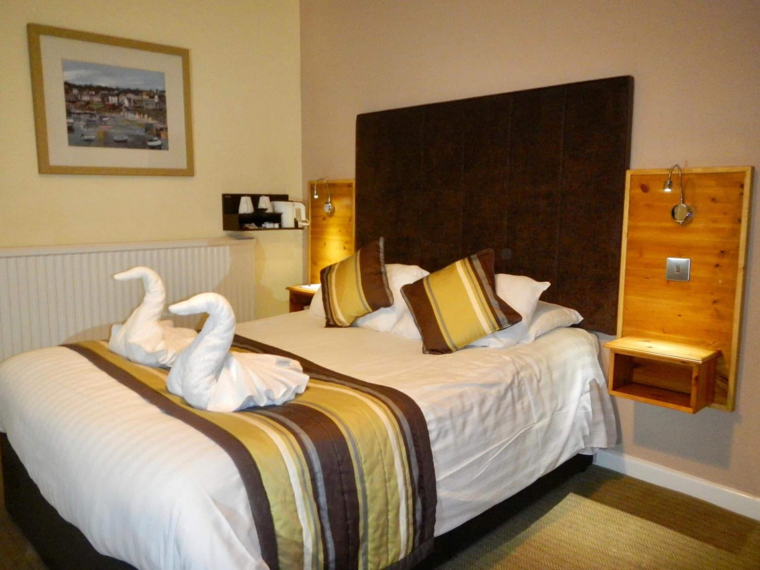 A hotel room with a large bed, two pillows, a headboard, a bedside table, a nightstand, a framed picture on the wall, and a radiator.