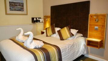 A hotel room with a large bed, two pillows, a headboard, a bedside table, a nightstand, a framed picture on the wall, and a radiator.